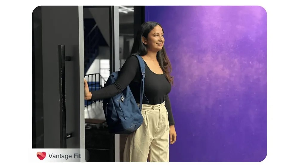 A woman standing near a doorway, smiling, wearing a black top and beige pants, with a backpack on her shoulder, against a purple background