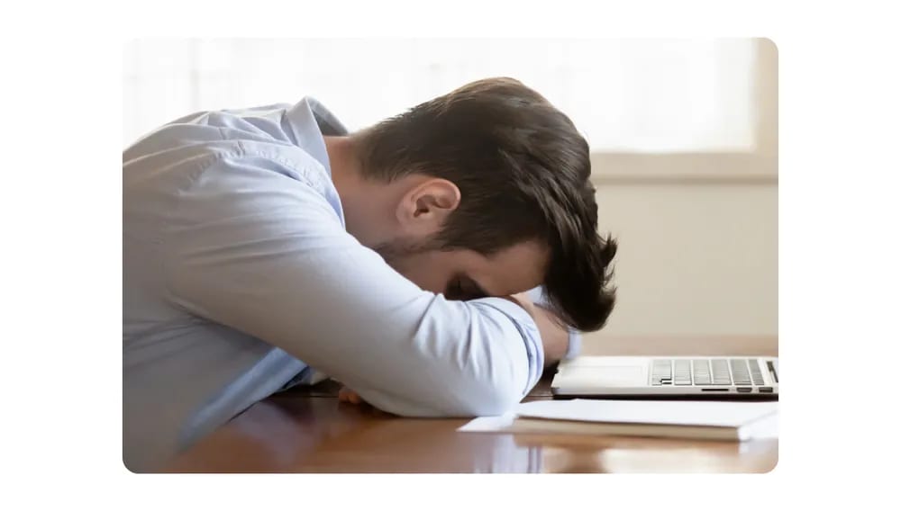 A tired employee napping at his desk with his head resting on folded arms near a laptop.png