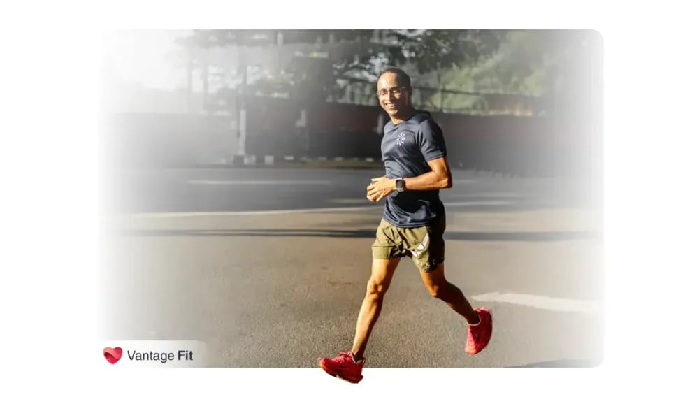 A man jogging outdoors, smiling while wearing a dark blue t-shirt, shorts, and red running shoes, with a watch on his wrist, captured in natural daylight