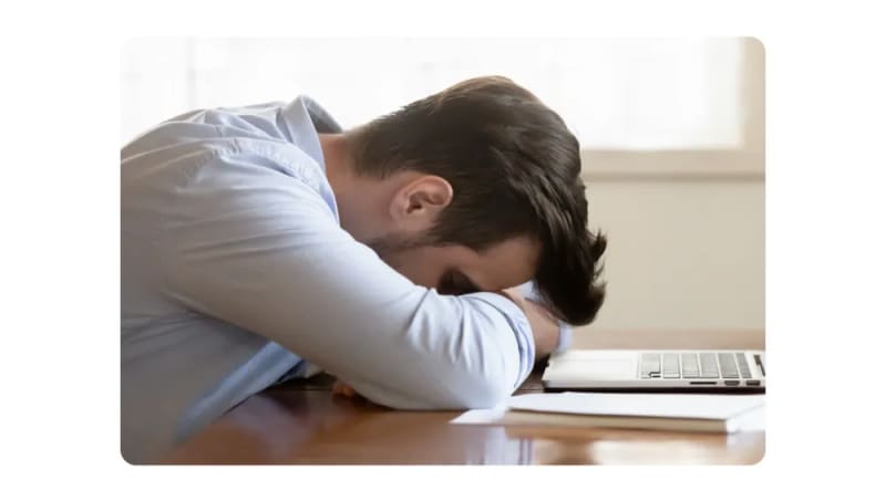 A tired employee napping at his desk with his head resting on folded arms near a laptop.png