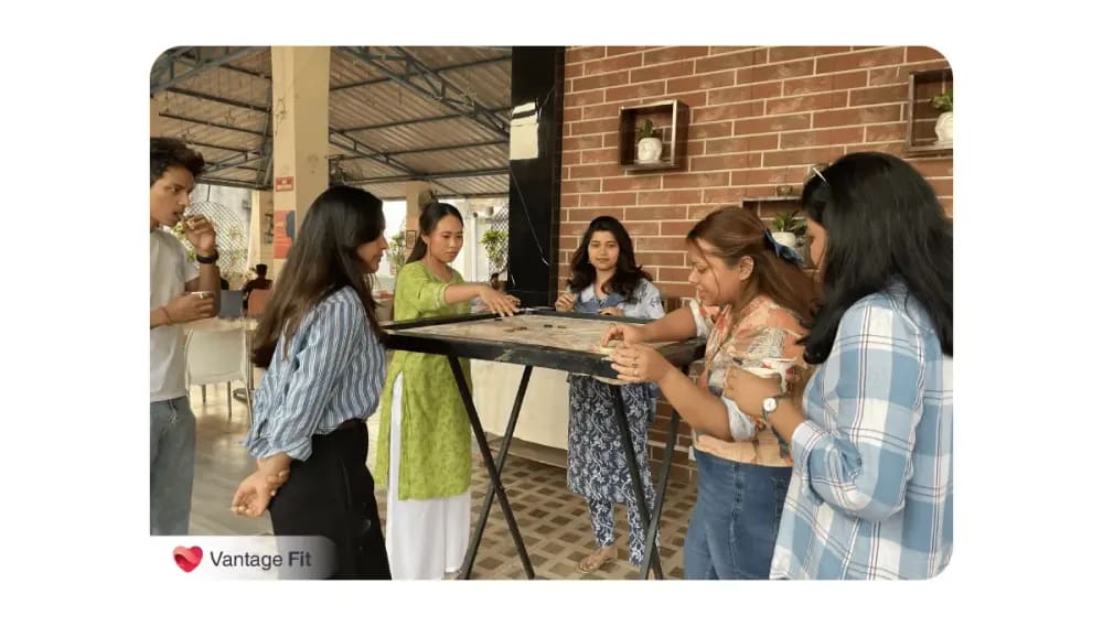 A group of colleagues, mostly women, gathered around a carrom board during a casual indoor break; some are playing while others watch and enjoy beverages, with Vantage Fit branding visible