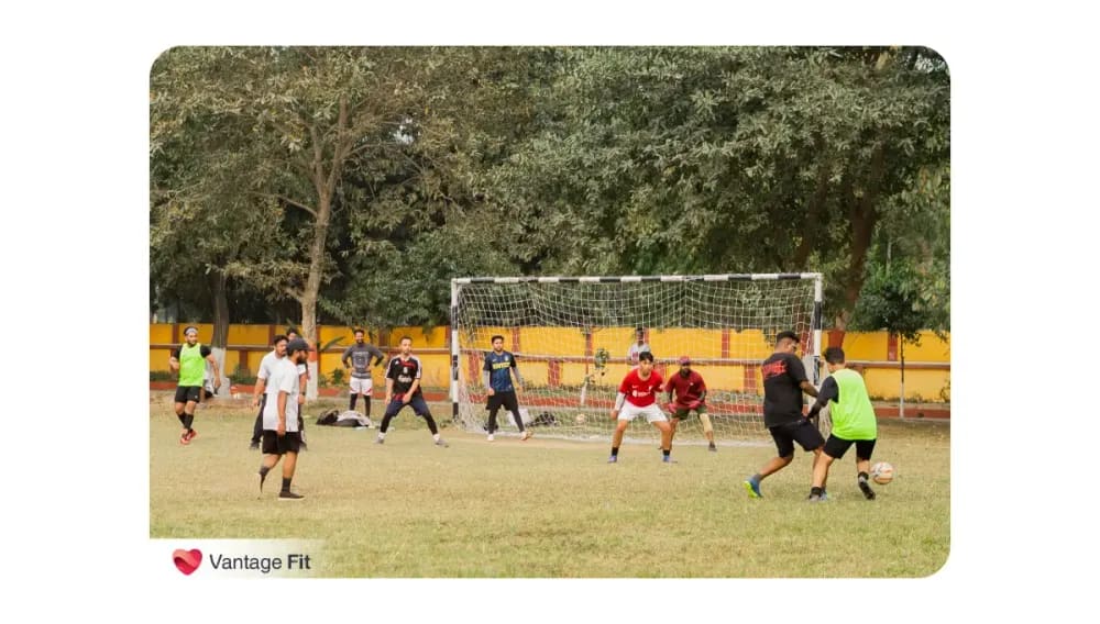 A group of men playing football on a grass field in front of a goalpost, some defending and others attacking, with the Vantage Fit watermark