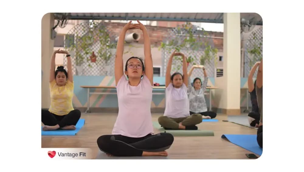 A group of women practicing seated yoga poses with arms stretched overhead on yoga mats in a peaceful indoor setting, branded with Vantage Fit