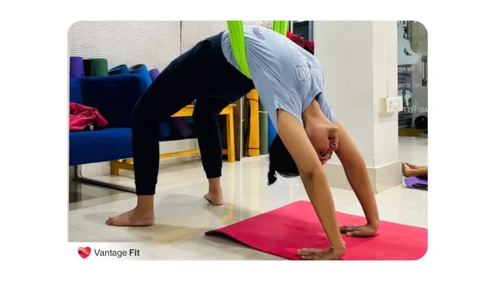 A woman practicing aerial yoga in a backbend pose supported by a hammock inside a well-lit fitness studio, showcasing flexibility and strength, with Vantage Fit branding