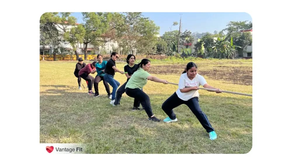 Eight women intensely focused in a tug-of-war challenge on a field, all pulling a thick rope with determination, featuring the Vantage Fit brand
